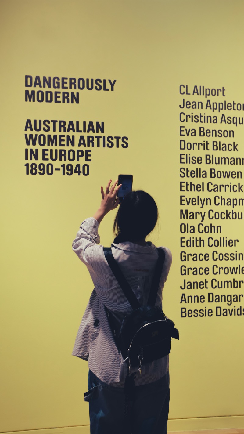 A gallery visitor, seen from behind, photographs the title wall of an exhibition on her phone