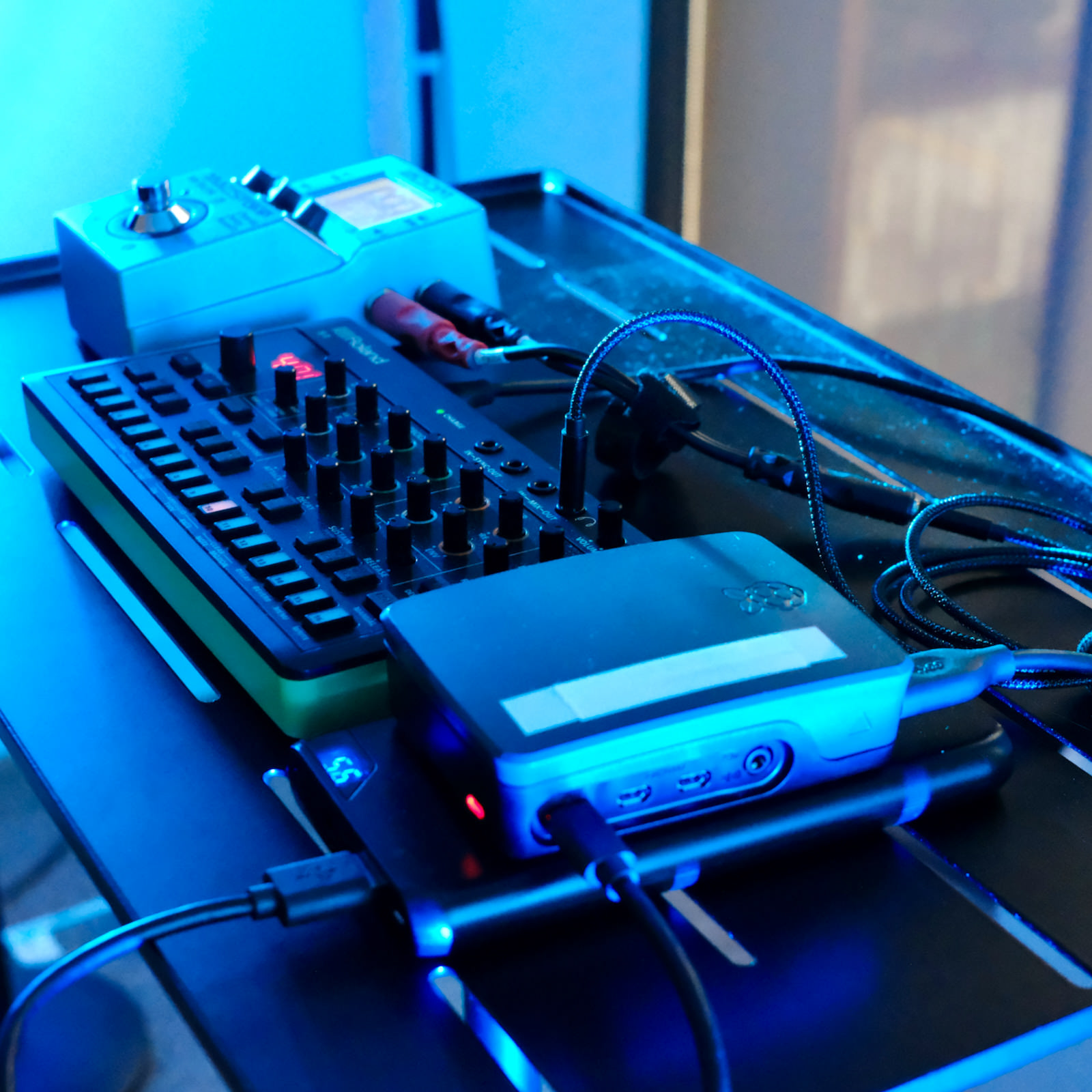 A Raspberry Pi in a black case sits in front of a small Roland S-1 synthesiser on a stage table, lit blue.