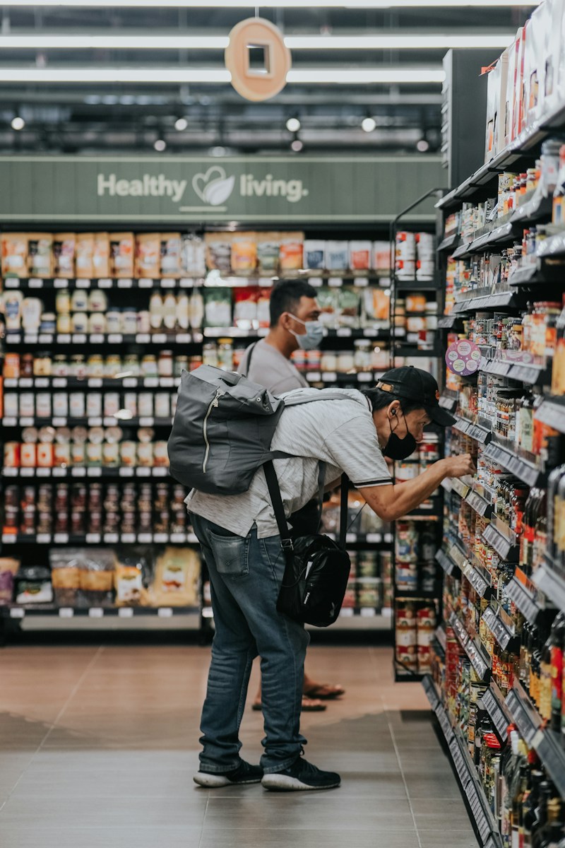 A masked shopper reaches into a densely stocked supermarket shelf under a Healthy living sign